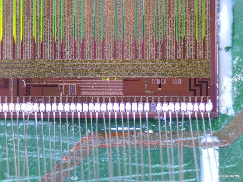 Microscope view of a chip edge: a row of aluminium bond pads, each with a wedge bond and a thin wire leaving downward toward the PCB; the chip's top metal routing visible above the pad row