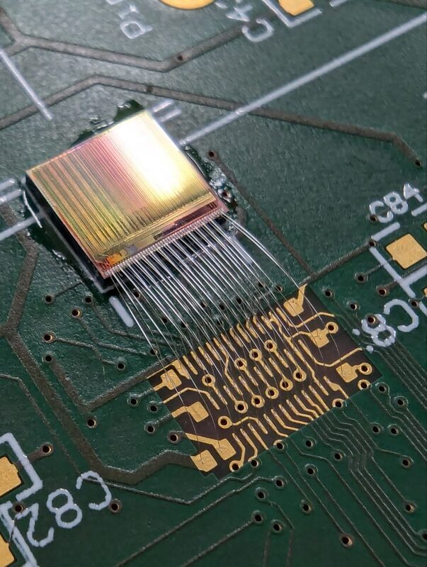 A bare die glued onto a PCB, gold-colored top metal catching the light, with a fan of thin bond wires running from the chip edge down to a field of bond fingers on the board