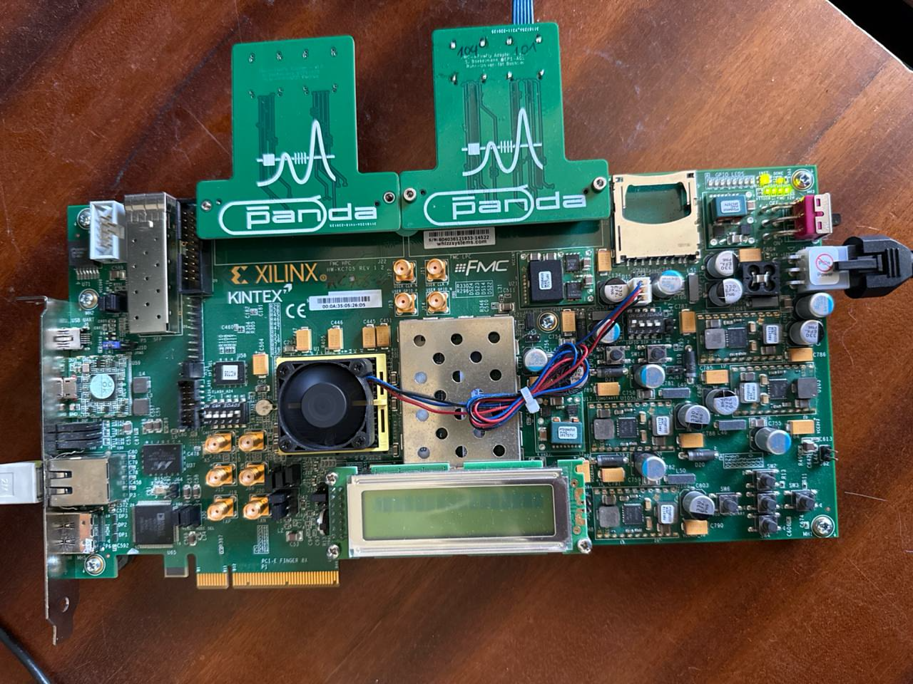A Xilinx Kintex-7 evaluation board in PCIe form factor, with two Panda FMC mezzanine cards attached, an SFP cage, SMA connectors, and a small cooling fan over the FPGA package