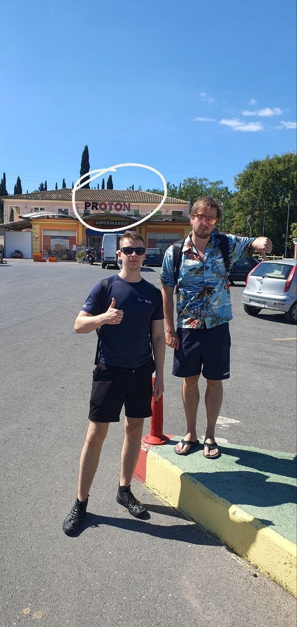 Stephan Bökelmann a.k.a MaxClerkwell and another researcher from RUB standing in a supermarket parking lot on Corfu — the store sign reads "PROTON". Stephan holds his thumb down, because he is firmly on Team Antiproton.